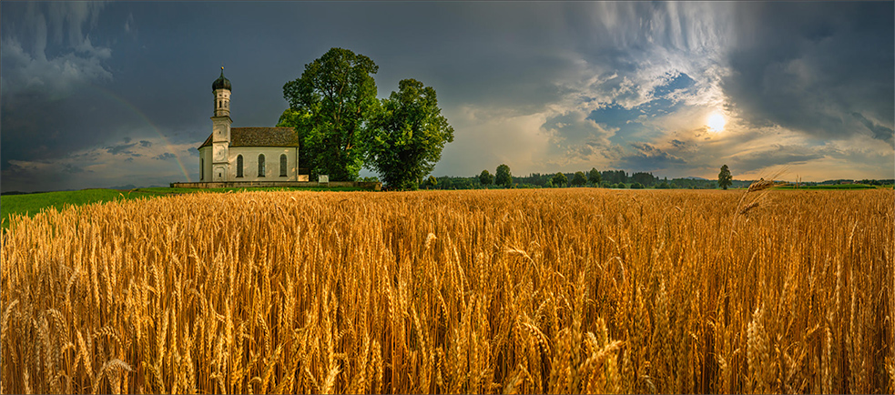 A beautiful picture of a church in Bavaria -- with wheat in the foreground and the sun and a rainbow in the background